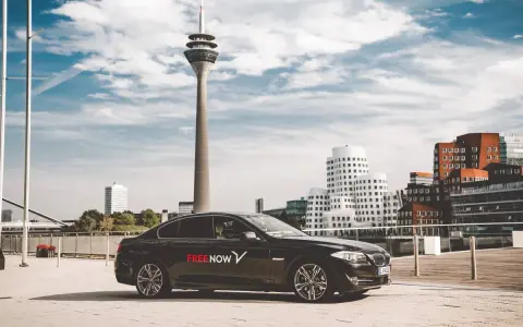 Schwarzer Wagen mit FREE NOW Logo steht vor dem Rheinturm in Düsseldorf. Ein FREE NOW Fahrer wartet auf seine Fahrgäste.