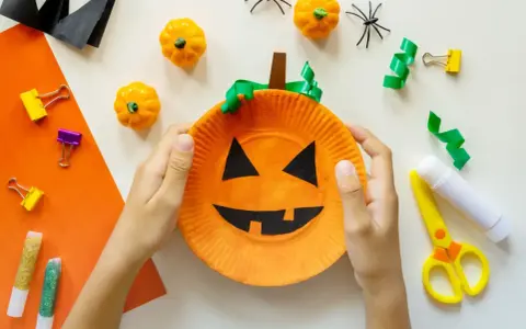 Barn gör halloween pyssel med en pumpa av papper. Små pumpor, spindlar och glitterpennor ligger redo.