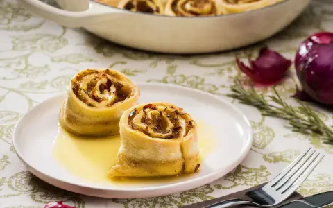 Zwei goldbraune Krautkrapfen im Sud auf einem Teller, im Hintergrund ein Auflauf mit weiteren Krapfen, Zwiebeln und Rosmarin. Ein leckeres Krautkrapfen Rezept.