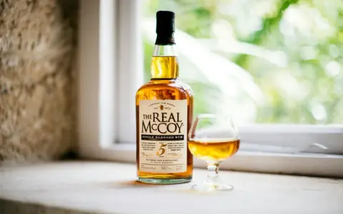 Ein besonderer Rum, "The Real McCoy", steht neben einem Glas mit bernsteinfarbener Flüssigkeit vor einem Fenster mit grünem Bokeh.