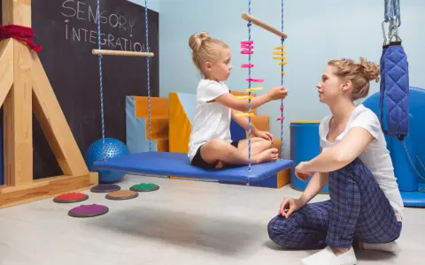 Dziecko siedzi na huśtawce w sali integracji sensorycznej, obok siedzi terapeuta.
