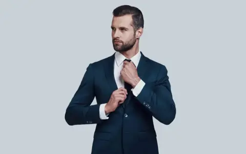 Elegancki mężczyzna w granatowym garniturze i białej koszuli poprawia krawat. Idealna stylizacja, gdy zastanawiasz się, jaka koszula do granatowego garnituru.