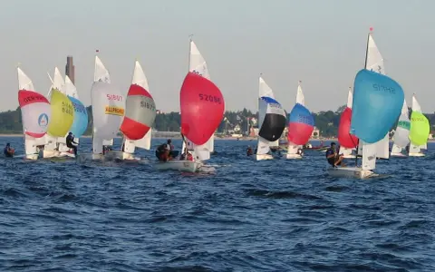 Bunte Segel dominieren das Bild einer spannenden Regatta. Mehrere kleine Boote mit Seglern sind auf dem Wasser unterwegs.