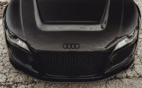 Przód czarnego Audi z maską z włókna węglowego. Widoczne reflektory, atrapa chłodnicy i logo marki.