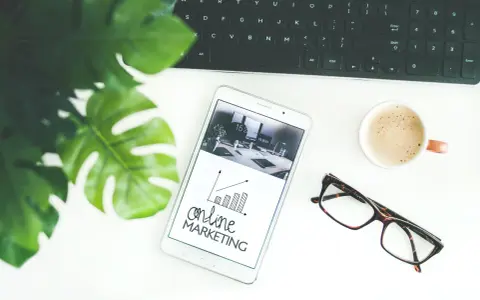 Biurko z tabletem pokazującym wykresy i hasło "online MARKETING", obok klawiatura, kawa i okulary. Idealne do social media studia.
