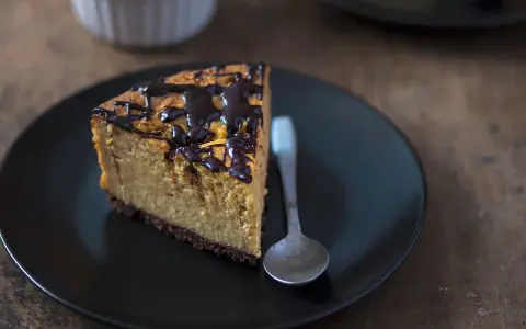 Tranche de cheesecake sans gluten sans lactose, nappée de chocolat noir, sur une assiette noire avec une cuillère.