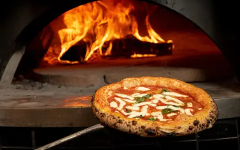 En perfekt italiensk pizza med mozzarella och basilika tas ur en vedeldad ugn med flammande eld.