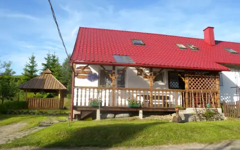 Uroczy domek z czerwoną dachem i drewnianą werandą, idealny na noclegi blisko Tarnicy. Obok altana i zielony ogród.