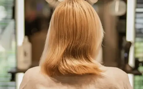 Blond włosy po zabiegu farba demi permanentna, delikatnie pofalowane, w tle rozmyte światła salonu fryzjerskiego.