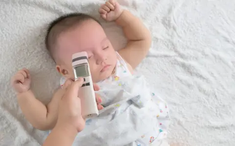 Jak mierzyć temperaturę u niemowlaka termometrem bezdotykowym? Ręka trzyma termometr przy czole śpiącego maluszka.