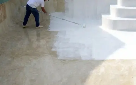 Hombre pintando una piscina de cemento con rodillo. La mitad de la piscina ya está pintada de blanco.