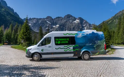 Biały bus Tatrzańskiego Parku Narodowego z grafiką górskiego krajobrazu, przypominającego Morskie Oko, stoi na kamiennej drodze.