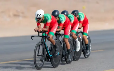Équipe cycliste marocaine en tenue rouge et verte, concentrée sur la course. Ils roulent en peloton, tels des pur sang arabes noirs, sur un parcours désertique.