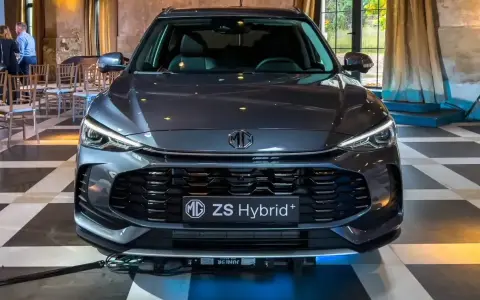 Szary MG ZS Hybrid+ prezentuje się elegancko na tle wnętrza z zasłonami i krzesłami.