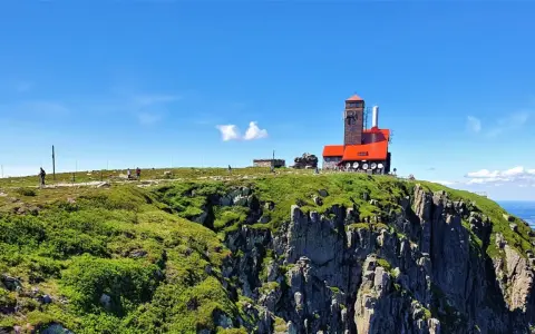 Widok na górskie **śnieżne kotły** z budynkiem obserwatorium na szczycie. Turyści spacerują po zielonych zboczach.