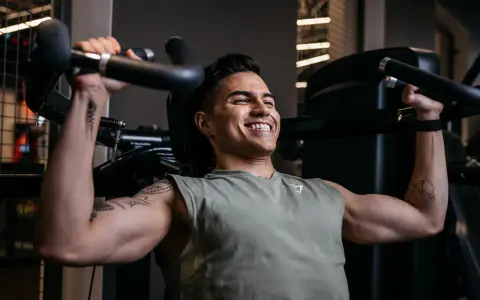 Ein Mann trainiert im Fitnessstudio, um seinen Brustmuskel aufzubauen. Er lächelt zufrieden, während er die Gewichte hebt.