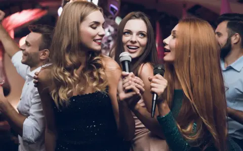 Trzy kobiety śpiewają do mikrofonów, bawiąc się przy piosenkach do śpiewania. W tle widać innych ludzi na imprezie.