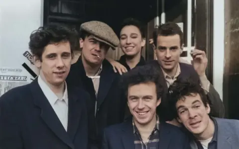 Medlemmar av the Pogues poserar glatt utomhus, några med cigaretter.