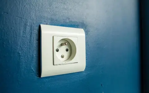 Prise électrique murale blanche sur fond bleu. Le branchement prise de terre est visible, prêt à connecter un appareil.