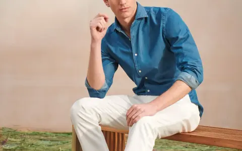 A man poses in a blue denim shirt and crisp white jeans, looking relaxed on a wooden bench.