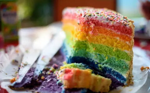 Ein Stück bunter **Regenbogen Kuchen** mit bunten Streuseln, das auf einem Teller liegt. Daneben liegen Krümel und ein Messer.