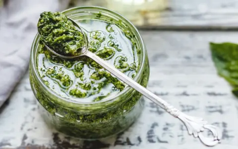 Un vasetto di pesto genovese fatto in casa con frullatore, pronto per essere gustato. Un cucchiaino d'argento raccoglie un po' di questa delizia verde.