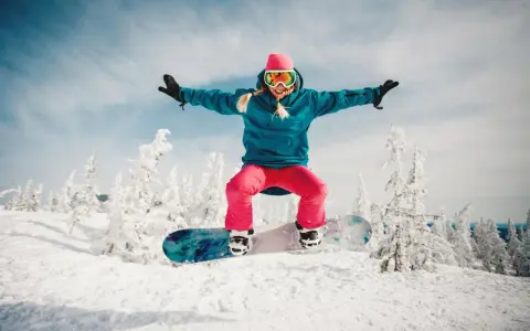Dziewczyna w różowej czapce i goglach skacze na snowboardzie na tle zaśnieżonych drzew. To jest właśnie snowboard!