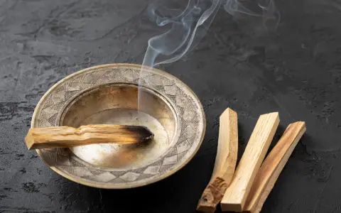 Un palo santo arde en un cuenco de metal, liberando humo blanco. A su lado, tres palos de palo santo sin quemar. Se puede quemar palo santo de noche para purificar el ambiente.