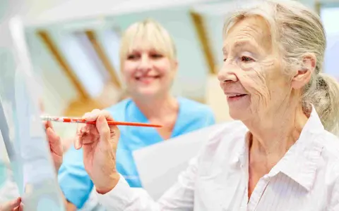 Anziana dipinge con un pennello, assistita da un'infermiera sorridente. Un esempio di terapie non farmacologiche per l'Alzheimer che stimolano la creatività.