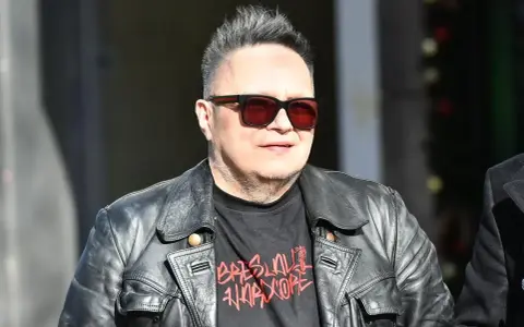 Charyzmatyczny wokalista w skórzanej kurtce i okularach przeciwsłonecznych, z charakterystyczną fryzurą.