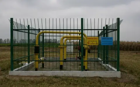 Żółty gazociąg wysokiego ciśnienia na działce, otoczony zielonym ogrodzeniem. Tablice ostrzegają przed gazem i ogniem.