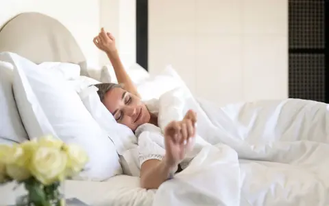 Une femme se réveille, s'étirant dans un lit douillet. La bonne literie blanche et les oreillers moelleux invitent à la détente.