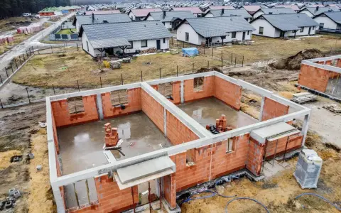 Budowa osiedla domów jednorodzinnych. Fundamenty i ściany parteru wznoszone z cegły, w tle gotowe domy. To początek drogi, jak zostać deweloperem.