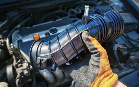 Gloved hand holds a black intake hose near a car engine. This part is often replaced during vacuum leak repair, affecting the vacuum leak repair cost.
