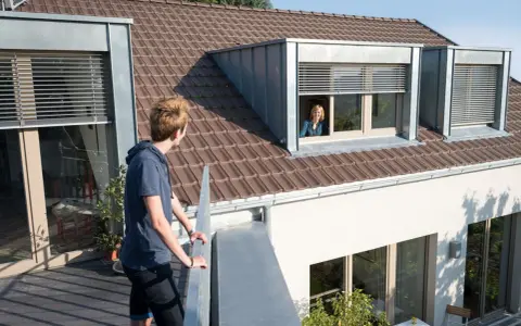Junge Person auf Balkon blickt zu Frau in Schleppgaube. Verschiedene Dachgauben Arten sind zu sehen.