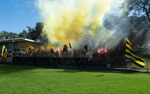 Fani Nielby Wągrowiec świętują, odpalając race i machając flagami. Żółto-czarne barwy dominują na trybunach.