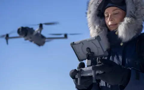 Woman in a fur-lined hood controls a flying drone in cold weather, looking at a tablet.