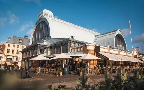 Människor sitter vid bord utomhus framför den imponerande **stora saluhallen** under en klarblå himmel.