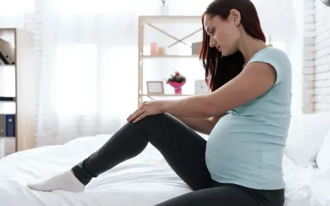 Schwangere Frau mit unruhigen Beinen, die auf einem Bett sitzt und ihr Bein berührt.