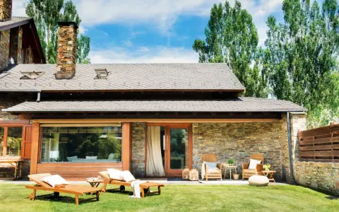 Fachadas de casas de pueblo modernas con piedra y madera, jardín con tumbonas y zona de estar exterior.