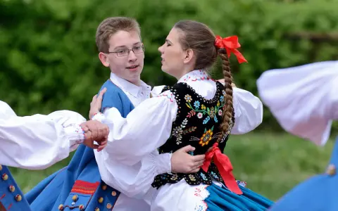 Młoda para w strojach ludowych tańczy mazura. Chłopiec w niebieskiej kamizelce, dziewczyna w haftowanej kamizelce i warkoczu z czerwoną wstążką.