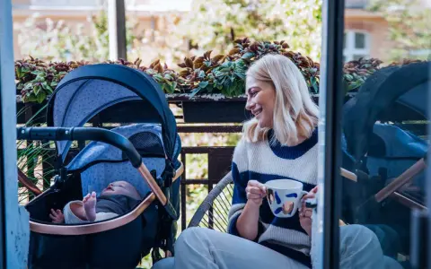 Uśmiechnięta mama z kawą na balkonie, obok wózek z dobrą amortyzacją i śpiące dziecko.