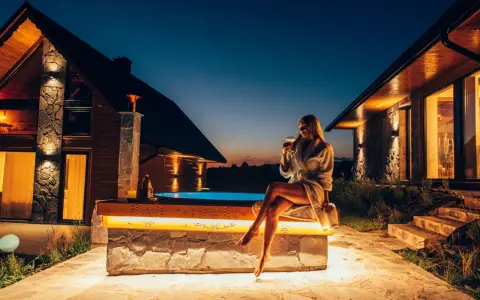 Kobieta w szlafroku z drinkiem siedzi przy jacuzzi przed górskim domkiem.