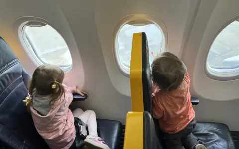 Dzieci patrzą przez okno samolotu Wizz Air. Dziewczynka w różowej bluzie i chłopiec w pomarańczowej koszulce, siedzą obok siebie.