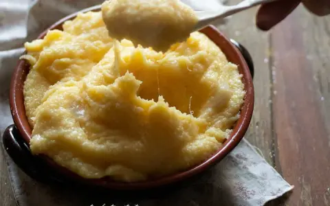Un cucchiaio solleva una porzione filante di polenta concia valdostana, calda e cremosa, in una ciotola di terracotta.