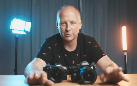 Mężczyzna z aparatami przed sobą, omawia podstawy fotografii. W tle niebieskie i pomarańczowe światła.