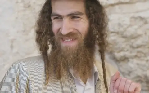 A smiling man with long, curly hair and a beard, wearing traditional Jewish attire. His appearance sparks curiosity about what ethnicity was Jesus.