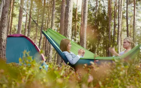 Två kvinnor njuter av en lugn stund i en hängmatta i skogen, med ett tält i närheten. En perfekt bild av campa Sverige.