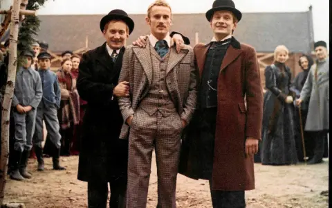 Trzech mężczyzn w eleganckich strojach, otoczonych tłumem, jakby w drodze do ziemi obiecanej.