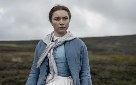 Florence Pugh i en blå klänning och jacka, med en scarf runt halsen, står i ett öppet landskap. En scen från filmer med Florence Pugh.
